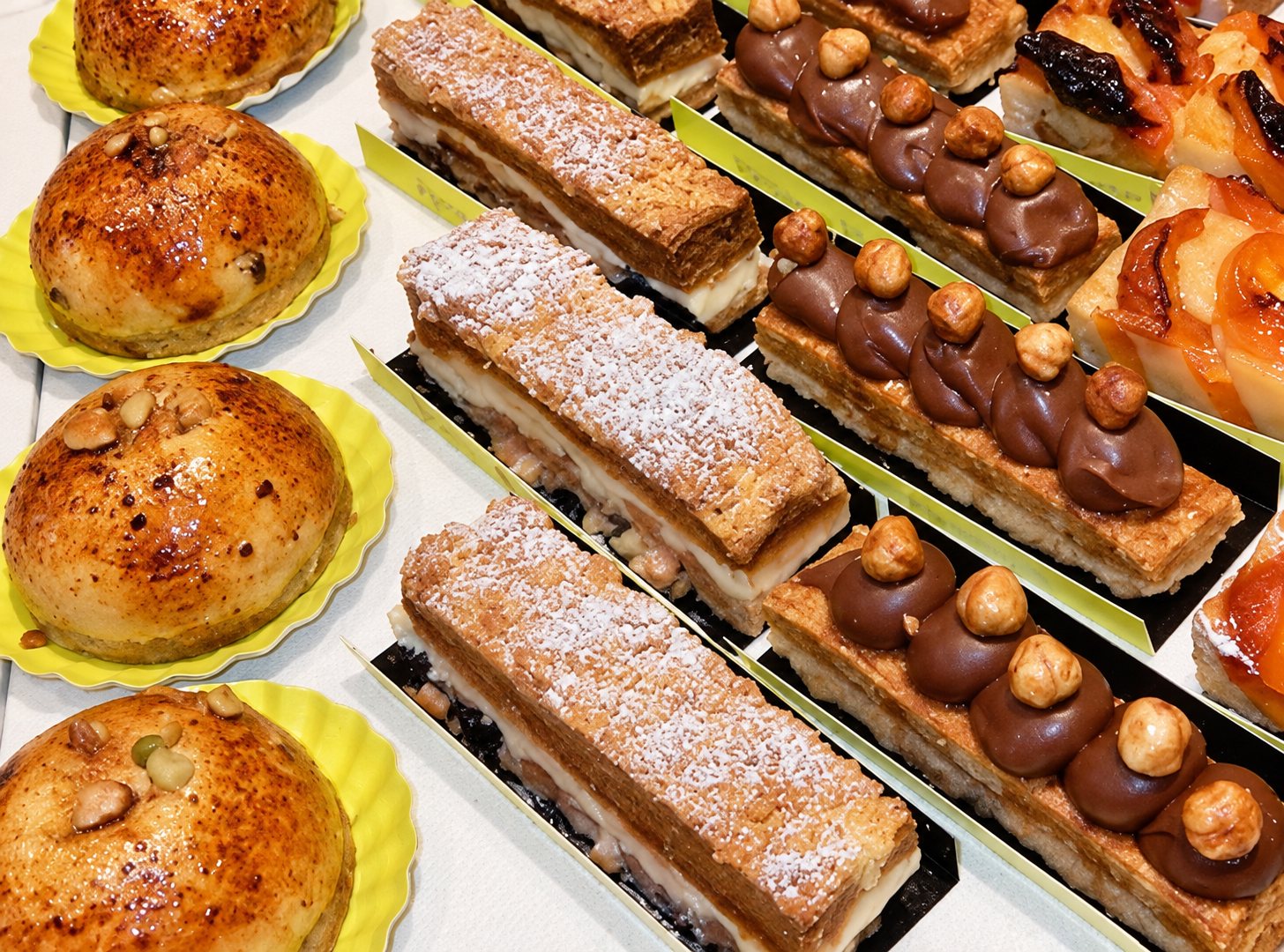 Vue de dessus de pâtisseries variées, brioches dorées et gâteaux décorés de noisettes, présentés sur des plateaux en vitrine.