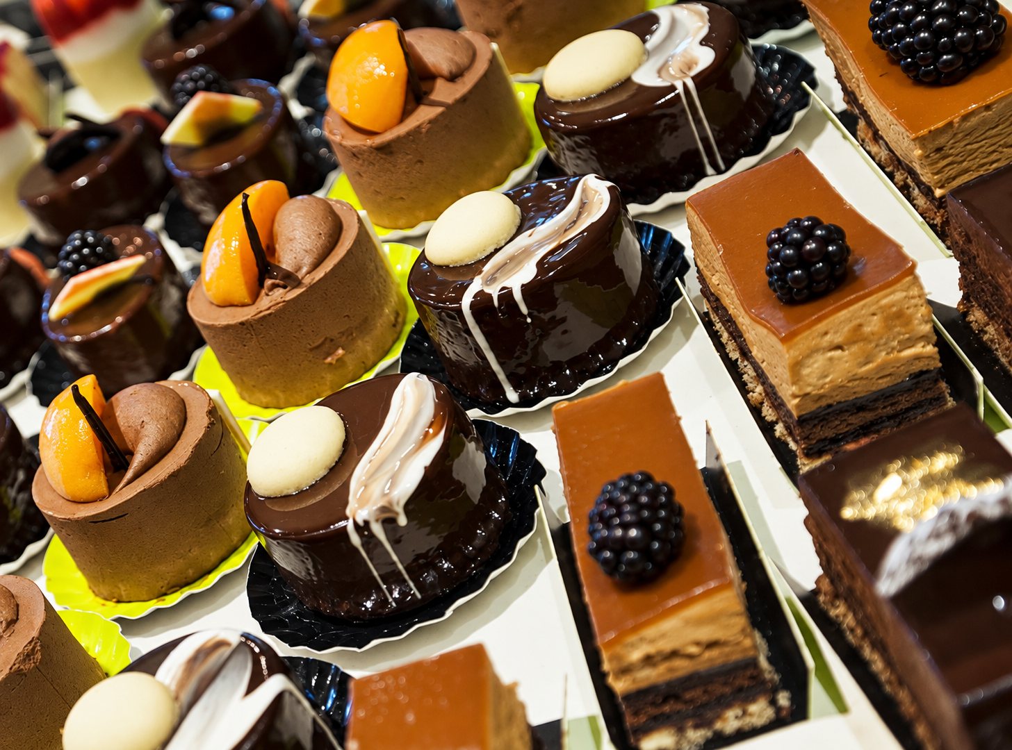 Rangées de petits gâteaux au chocolat, mousses à l'abricot, entremets surmontés de mûres et glaçages brillants, présentés sur des plateaux de pâtisserie.