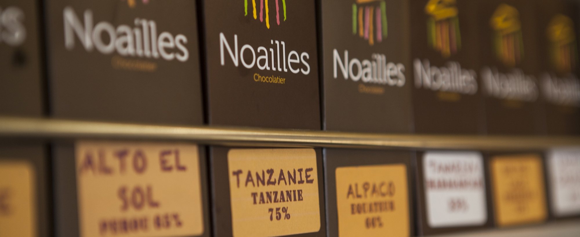 Rangée de boîtes marron de la chocolaterie Noailles sur une étagère, avec des étiquettes manuscrites indiquant l'origine du cacao, comme Tanzanie 75 %.