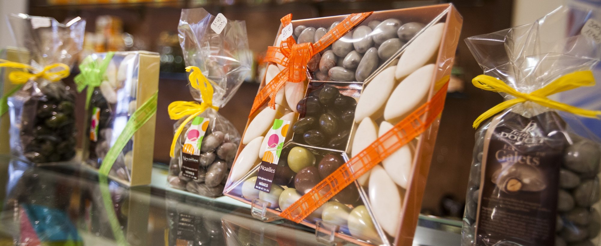 Sur une vitrine de boutique, des boîtes et sachets de dragées et de galets au chocolat sont décorés de rubans orange, jaunes et verts.