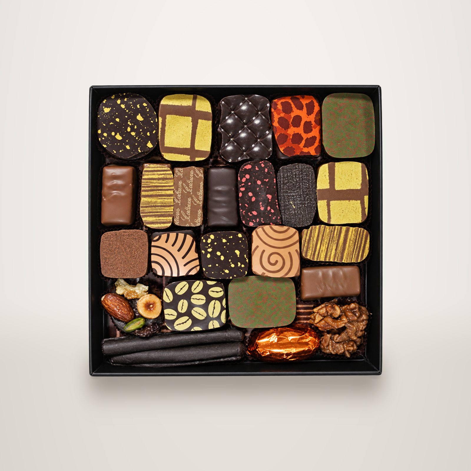 Vue de dessus d'un coffret noir contenant des chocolats artisanaux de formes, couleurs et décors variés, avec quelques noix et fruits secs.