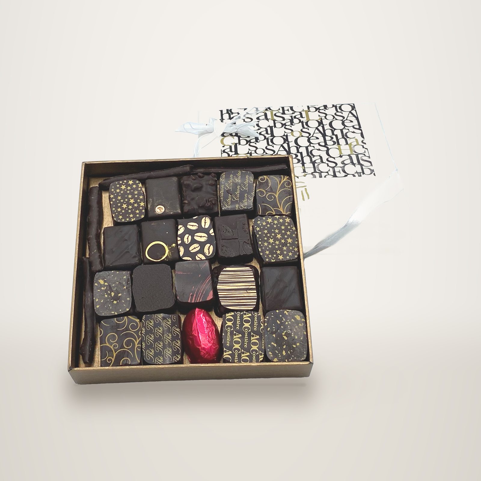 Un coffret doré ouvert contenant des chocolats noirs aux motifs dorés et un chocolat enveloppé de rouge, posé devant son couvercle blanc orné de lettres noires et d'un ruban.