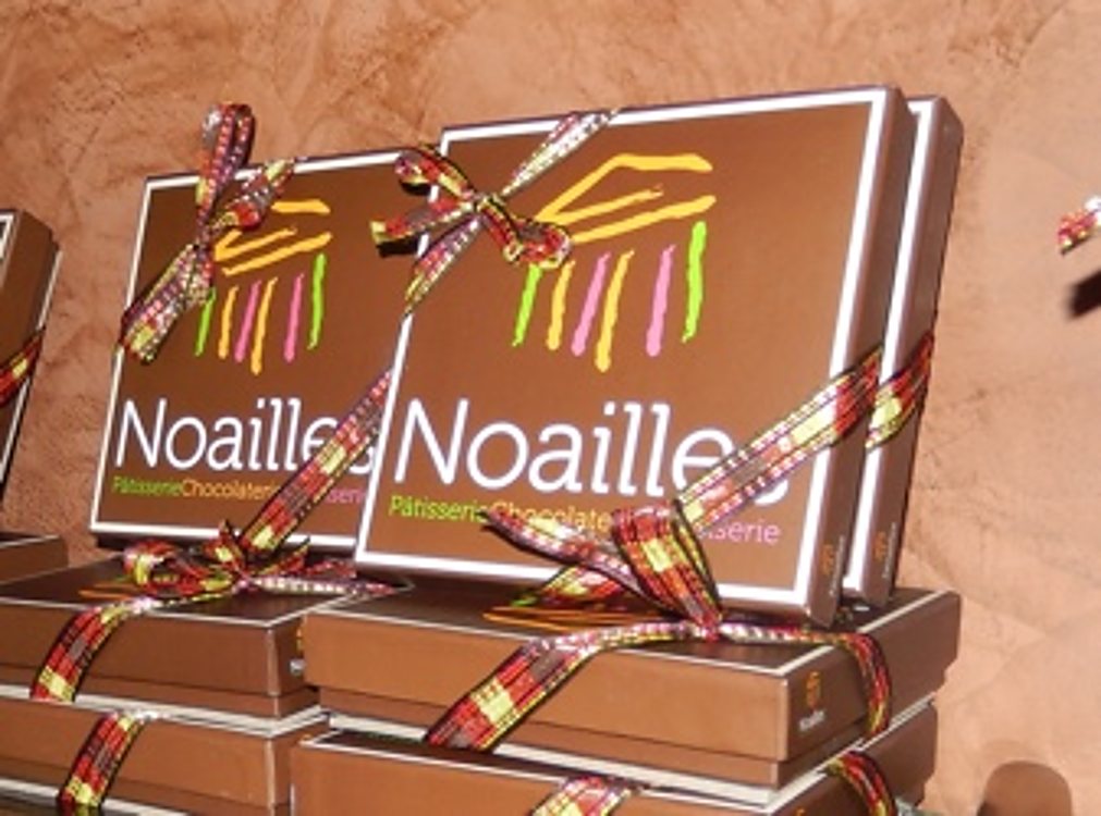 Des coffrets bruns de la marque Noaille, ornés de rubans à carreaux rouges et dorés, sont empilés devant un fond brun texturé.