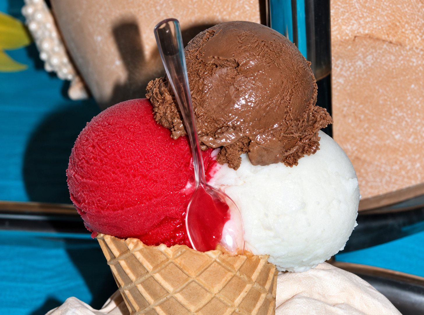 Un cornet gaufré garni de trois boules de glace, l'une rouge, l'une blanche et l'une au chocolat, avec une petite cuillère en plastique transparente.