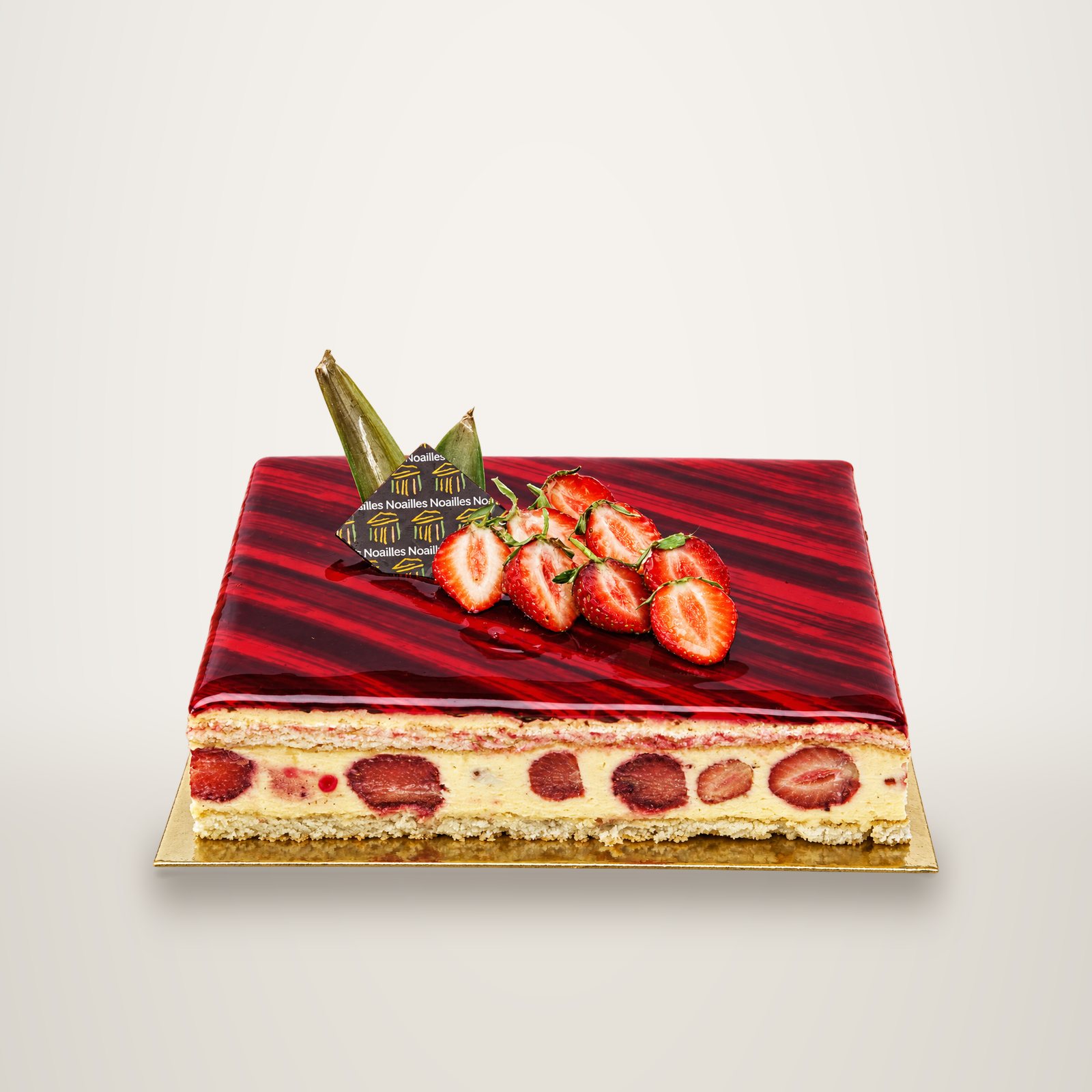 Un entremets rectangulaire aux fraises, nappé d'un glaçage rouge brillant, décoré de fraises coupées en deux et d'une plaque en chocolat portant le nom Noailles, posé sur un carton doré.