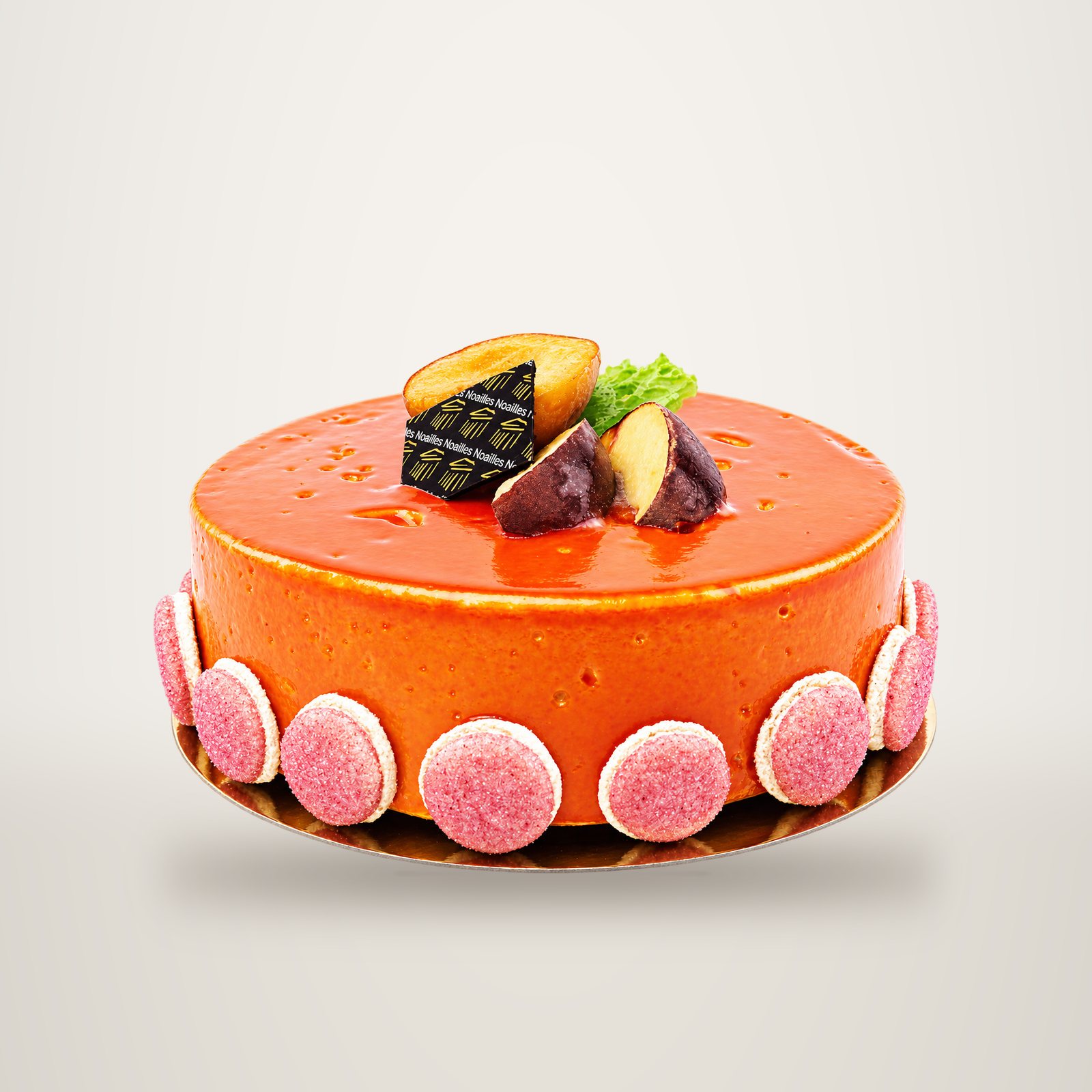 Un entremets au glaçage orange brillant, entouré de macarons roses et décoré de morceaux de marron et d'une plaque en chocolat portant le nom Noailles.