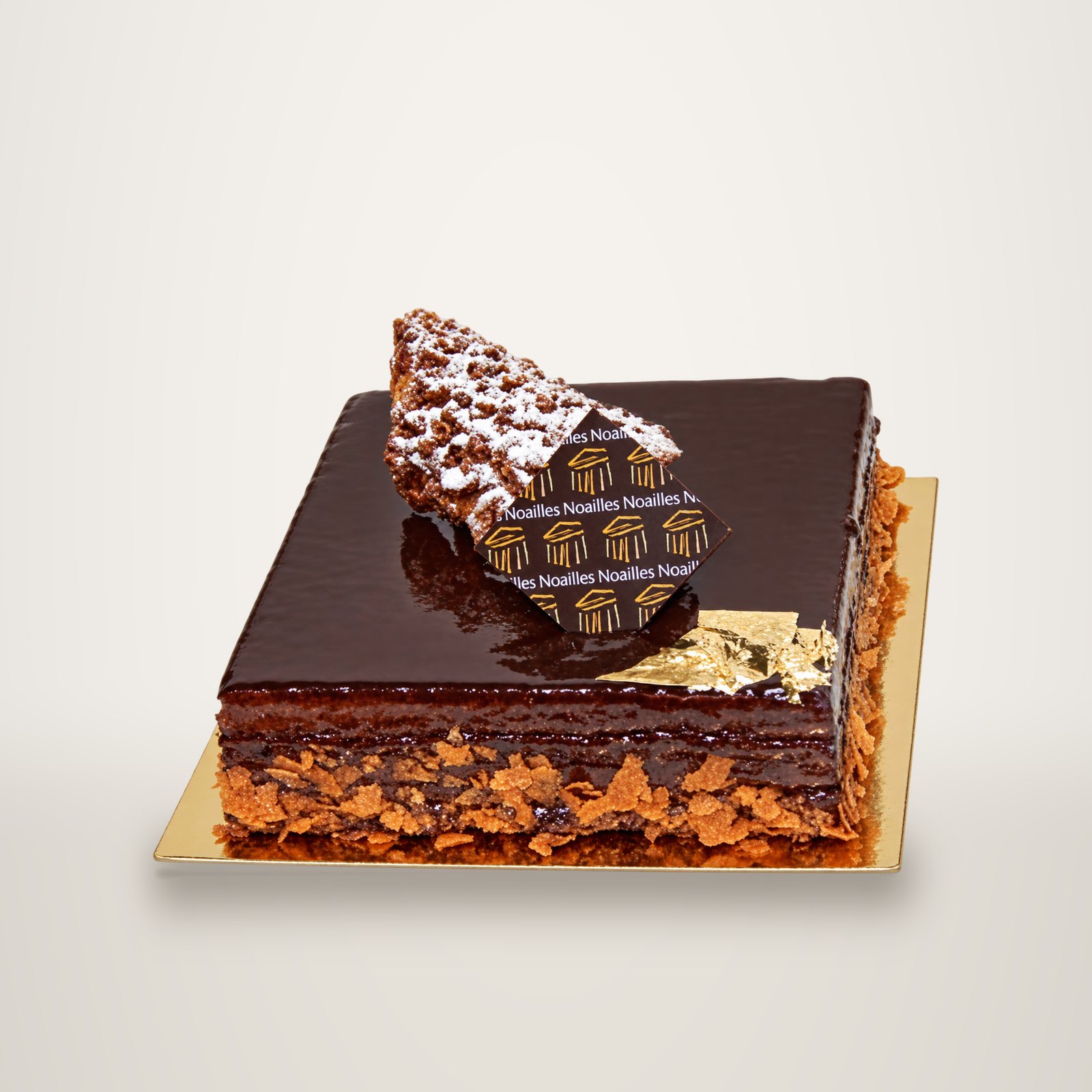 Un gâteau carré au chocolat à la surface laquée, décoré d'un éclat de praliné, d'une plaquette Noailles et de feuilles d'or, posé sur un carton doré.