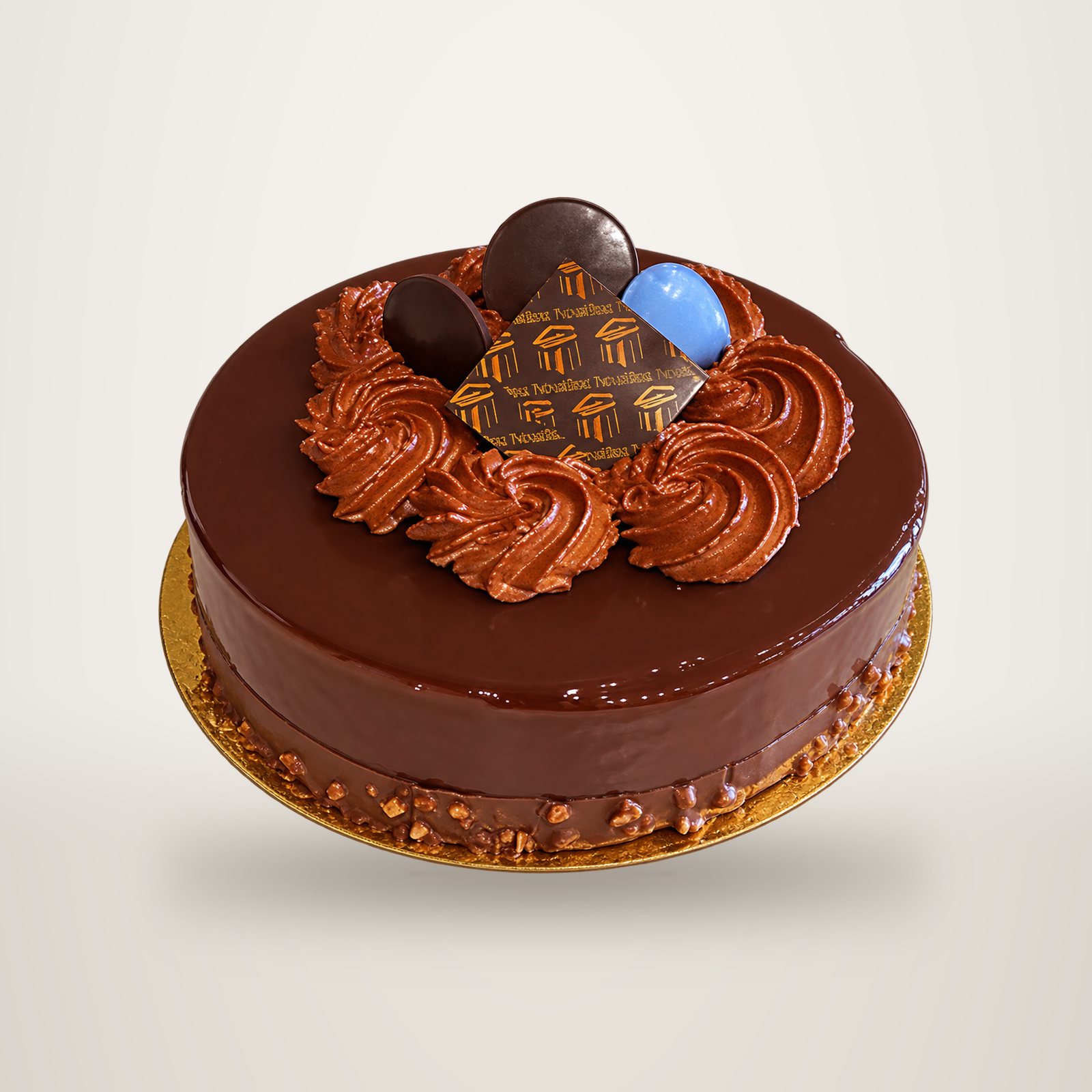 Un gâteau rond au chocolat à la surface brillante, décoré de rosettes de crème au chocolat, de disques en chocolat et d'une plaquette ornée d'un motif, posé sur un carton doré.