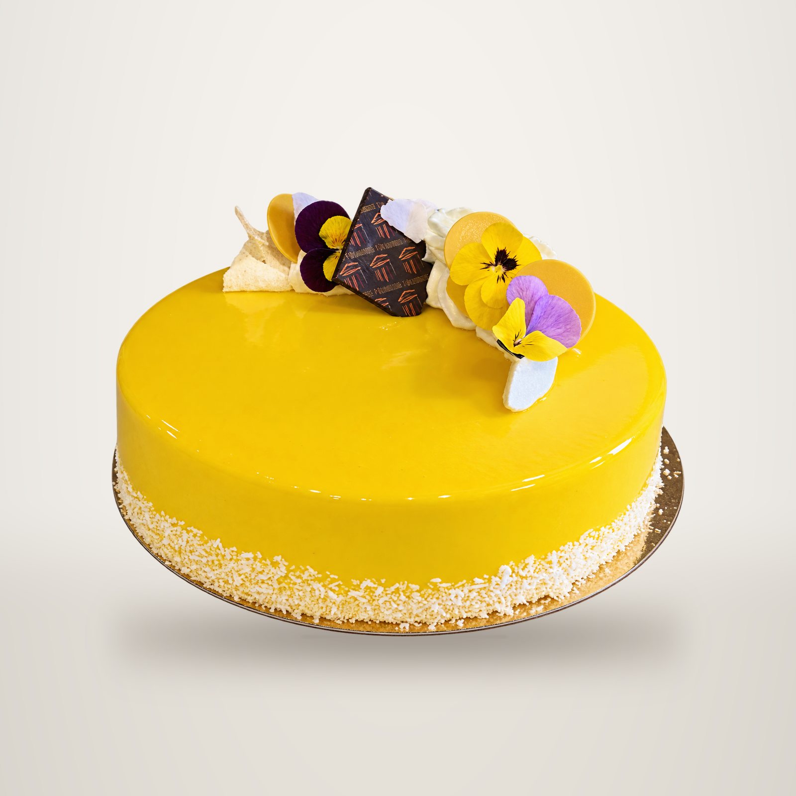 Un gâteau rond au glaçage miroir jaune vif, orné de pensées comestibles et d'un carré de chocolat, posé sur un support doré avec de la noix de coco sur le bord.