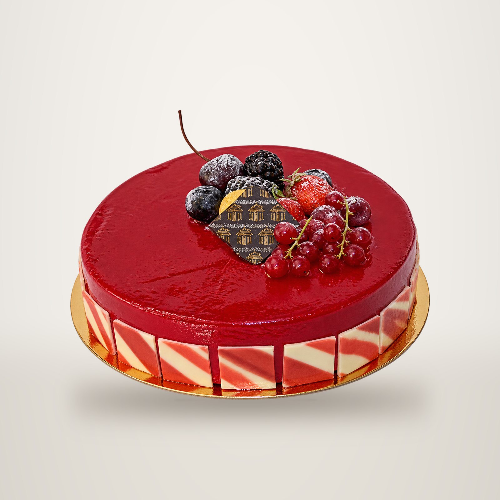 Un gâteau rond recouvert d'un glaçage rouge brillant, décoré de mûres, de groseilles et d'une plaque en chocolat, posé sur un support doré.