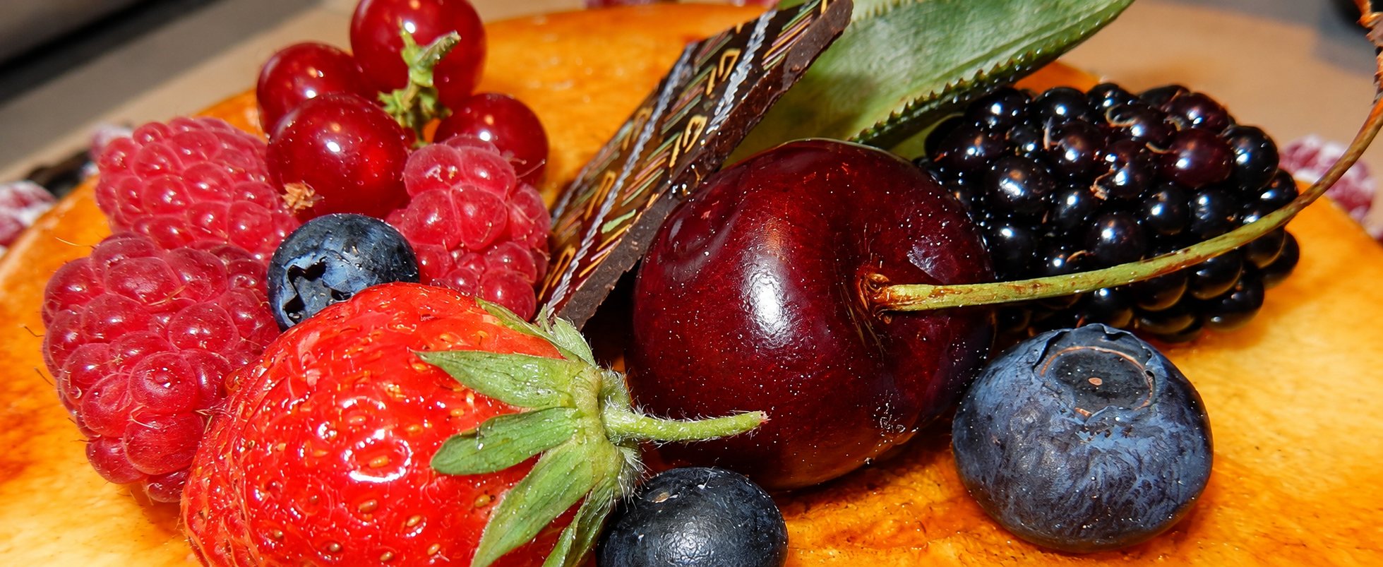 Gros plan sur un dessert nappé garni de framboises, d'une fraise, de myrtilles, d'une mûre, d'une cerise, de groseilles et d'une plaque de chocolat décorée.