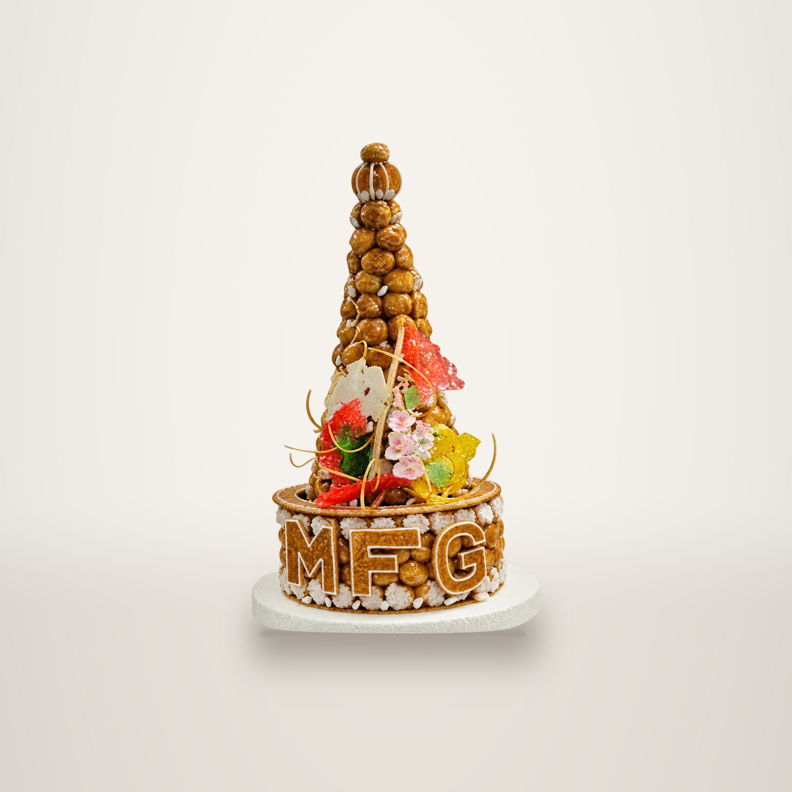 Une pièce montée en cône de choux caramélisés, décorée de fleurs en sucre colorées, avec les lettres MFG sur la base, photographiée sur fond blanc.