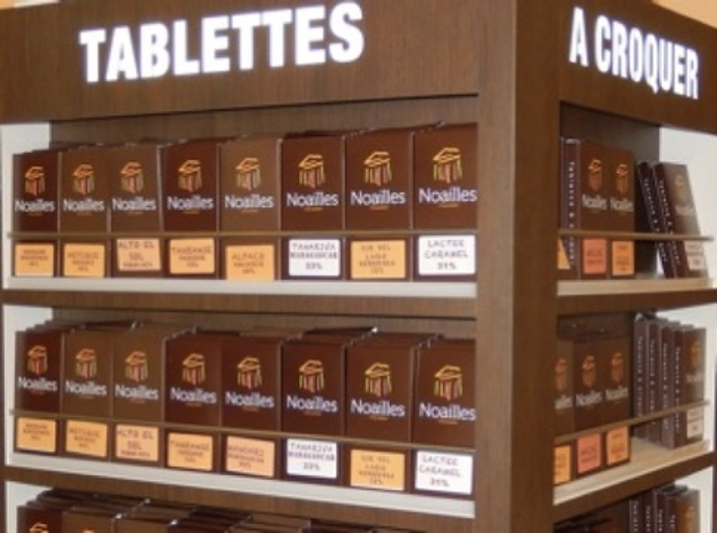 Un présentoir en bois sombre présente plusieurs rangées de tablettes de chocolat de la marque Noailles, avec des étiquettes de prix et de saveurs sous chaque paquet.