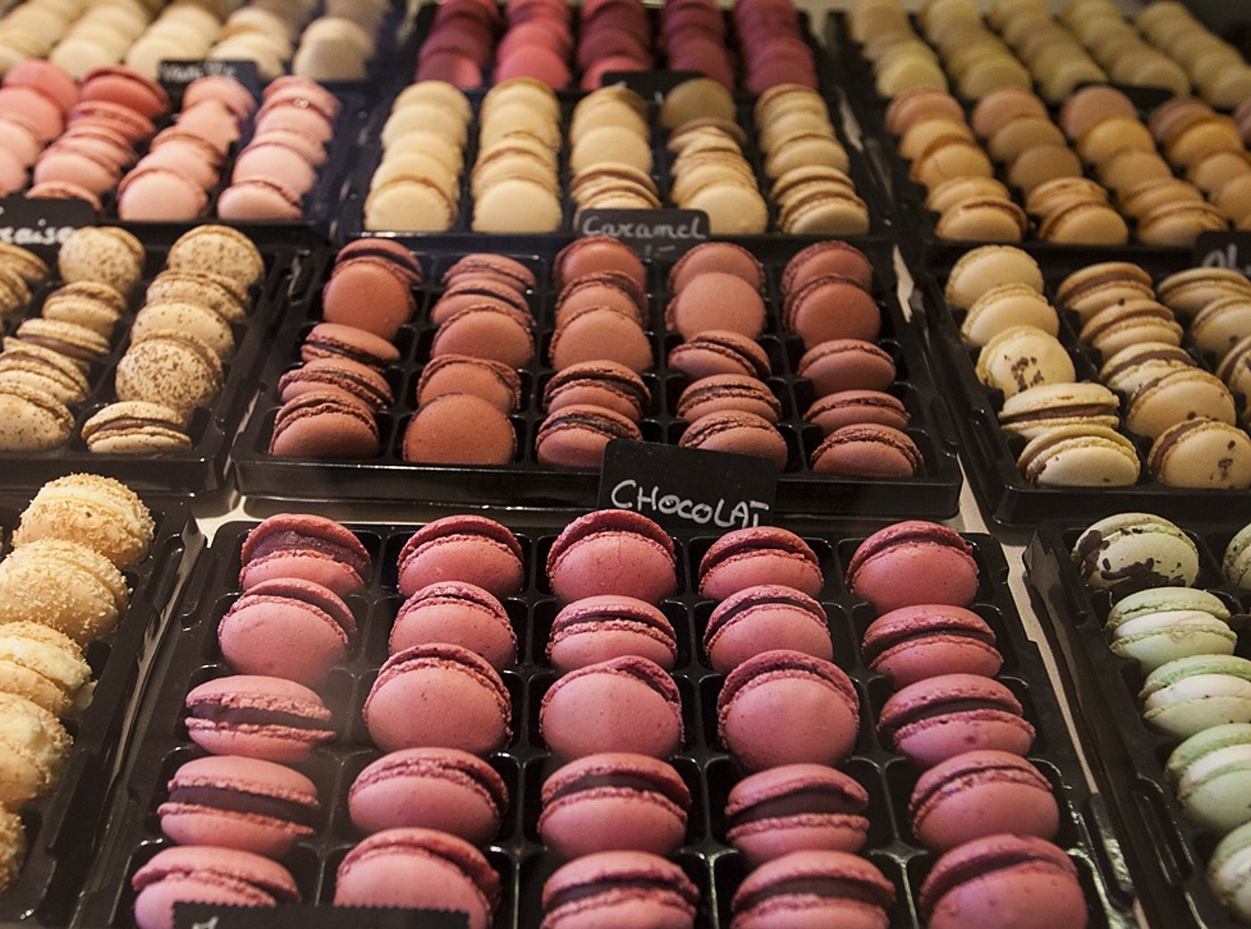Des rangées de macarons roses, beiges, marron et verts disposés dans des plateaux noirs avec des étiquettes à la craie, dont Caramel et Chocolat.