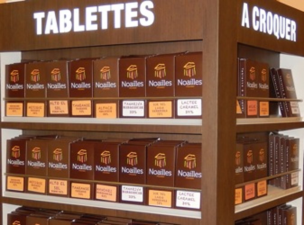 Un présentoir en bois sombre présente des rangées de tablettes de chocolat de la marque Noailles, sous une enseigne lumineuse « Tablettes » et « À croquer ».