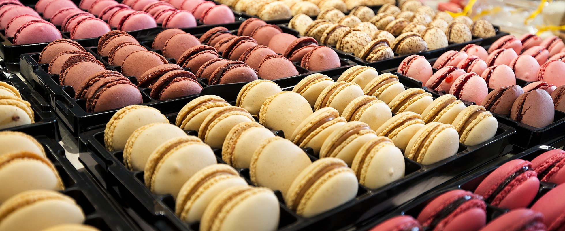 Plusieurs rangées de macarons beiges, rose et chocolat alignés dans des bacs noirs, présentés à la vente dans une pâtisserie.