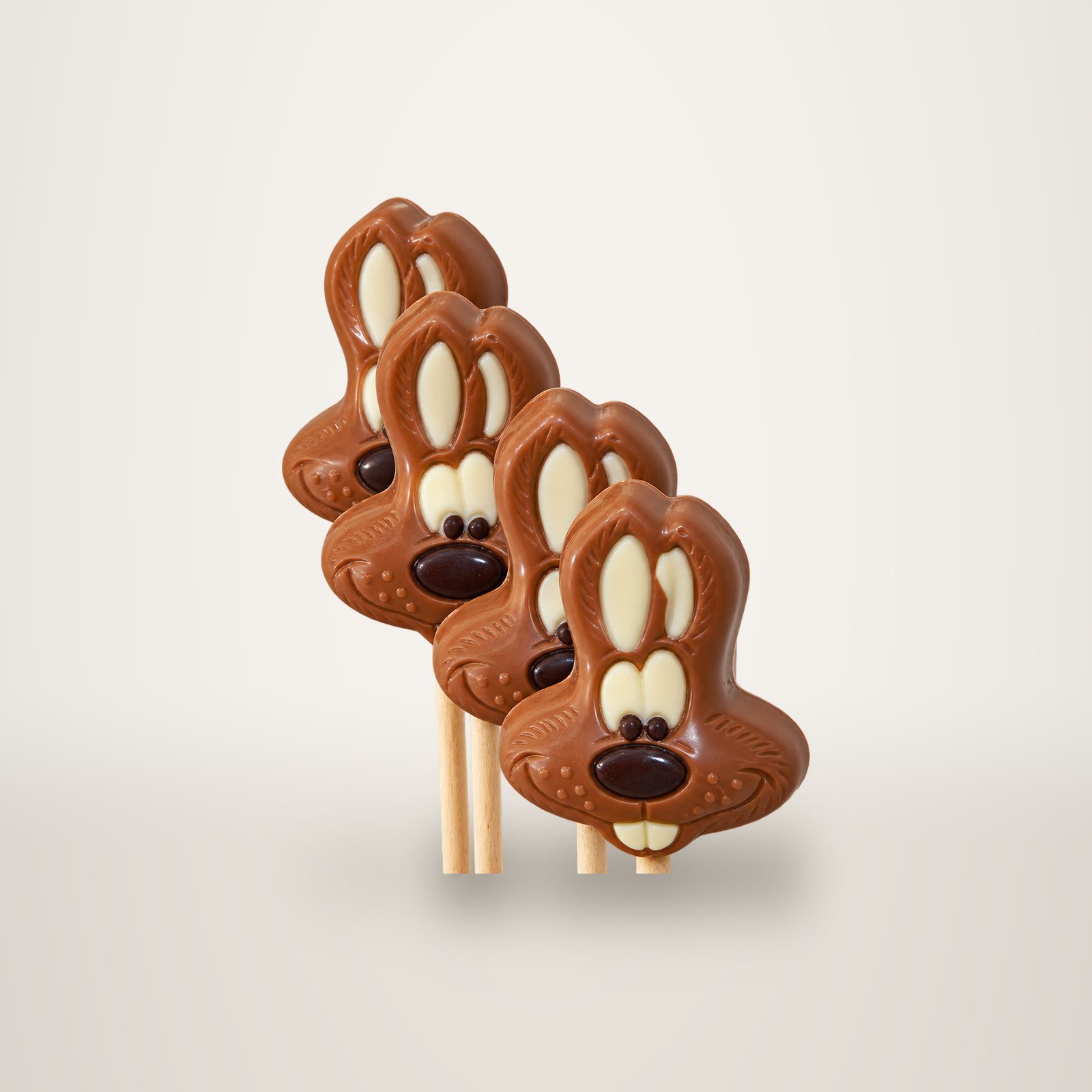 Quatre sucettes en chocolat au lait en forme de tête de lapin, avec museau et yeux en chocolat, posées sur des bâtonnets devant un mur clair.