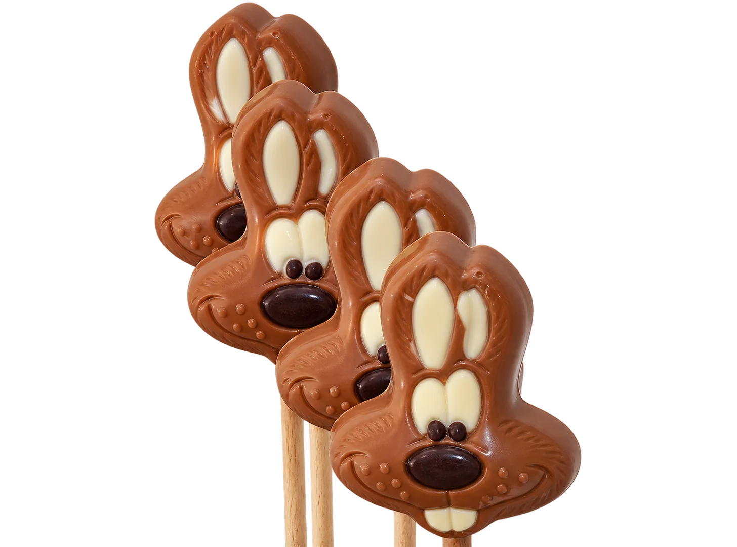 Quatre sucettes en chocolat au lait en forme de tête de lapin aux oreilles décorées, posées sur des bâtonnets devant un mur clair.