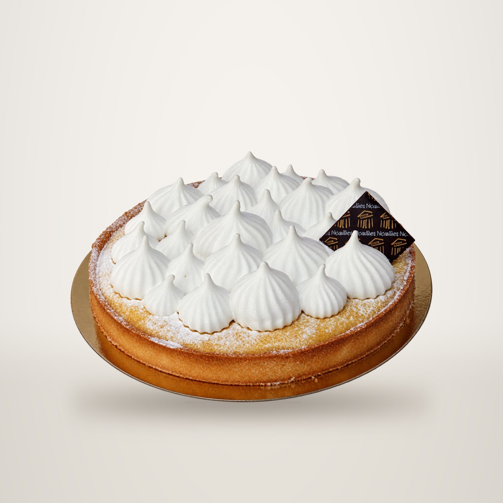 Une tarte au citron surmontée de pointes de meringue blanche, posée sur un carton doré, avec une petite plaque décorative sombre portant une inscription illisible.