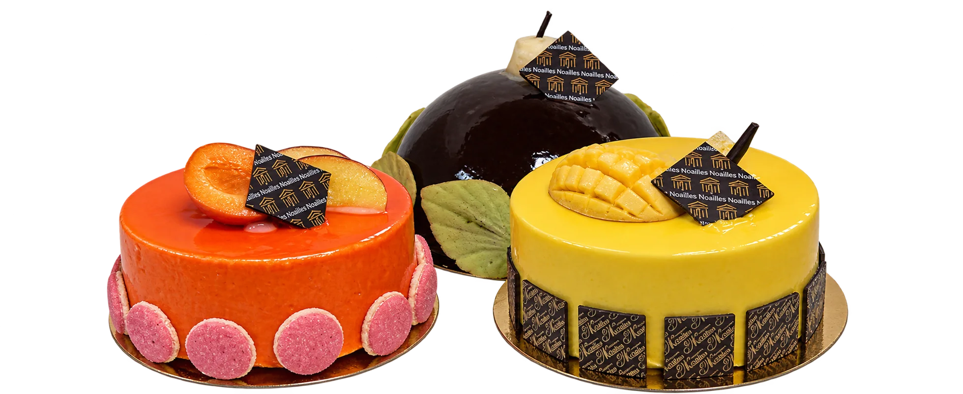 Trois gâteaux de pâtisserie : un entremets orange à l'abricot, un dôme au chocolat brillant et un entremets jaune à la mangue, posés devant un fond clair.