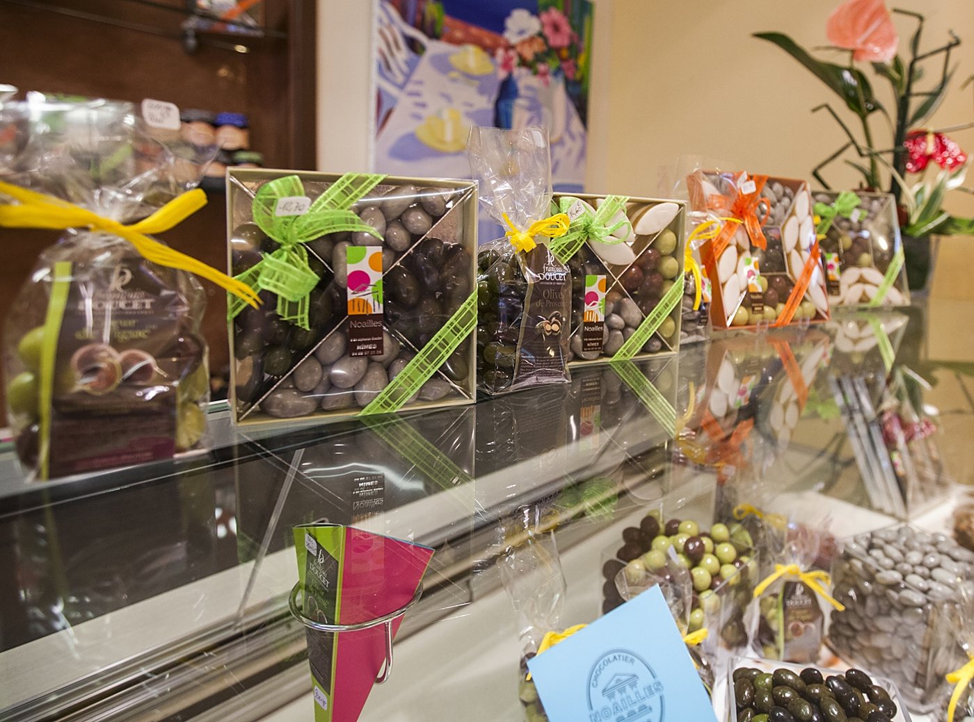 Une vitrine de chocolatier présente des coffrets et des sachets de chocolats et de dragées, ornés de rubans verts, jaunes et orange.