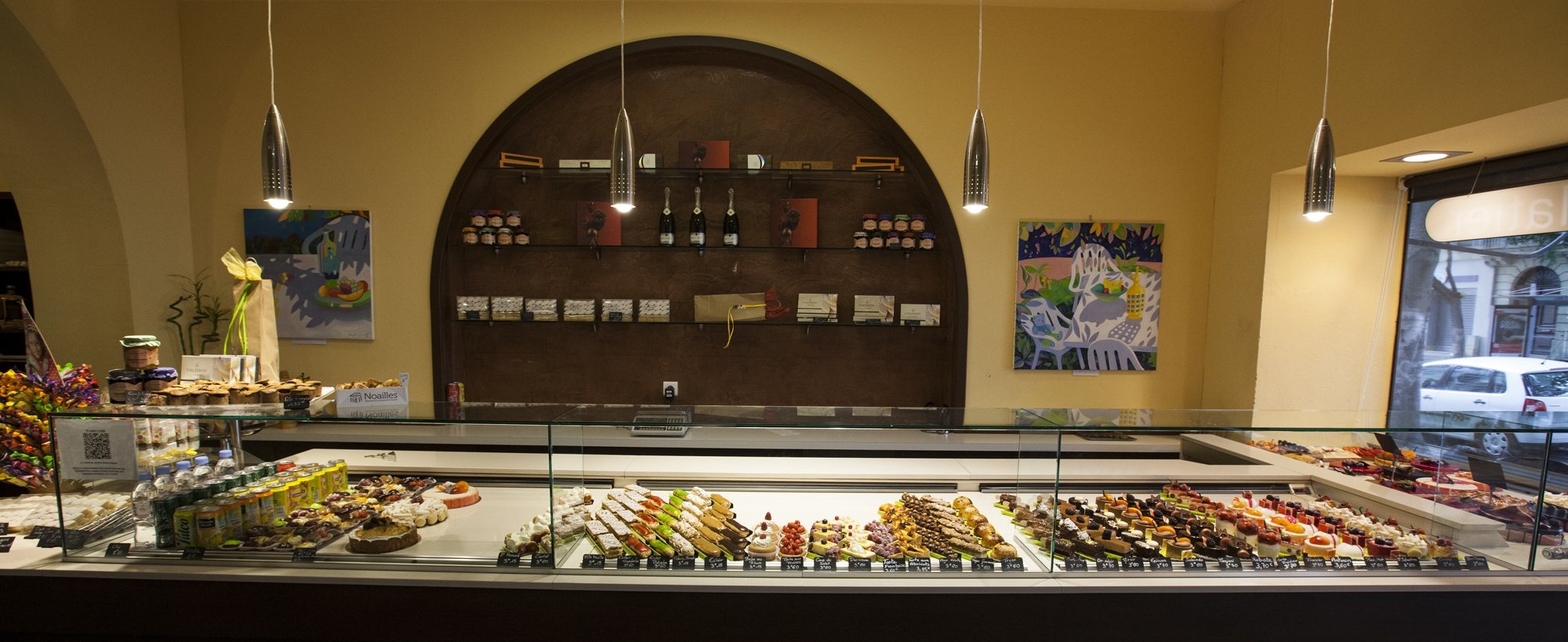 Intérieur d'une pâtisserie avec une longue vitrine remplie de gâteaux et de petits fours, devant une alcôve en arche garnie d'étagères de chocolats et de bouteilles.