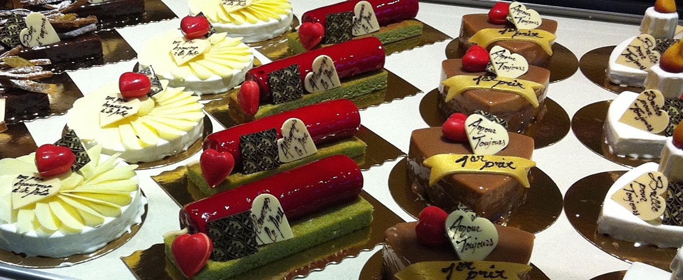 Des gâteaux individuels rouges, bruns et blancs décorés de cœurs en chocolat blanc portant des messages écrits à la main sont alignés dans une vitrine de pâtisserie.