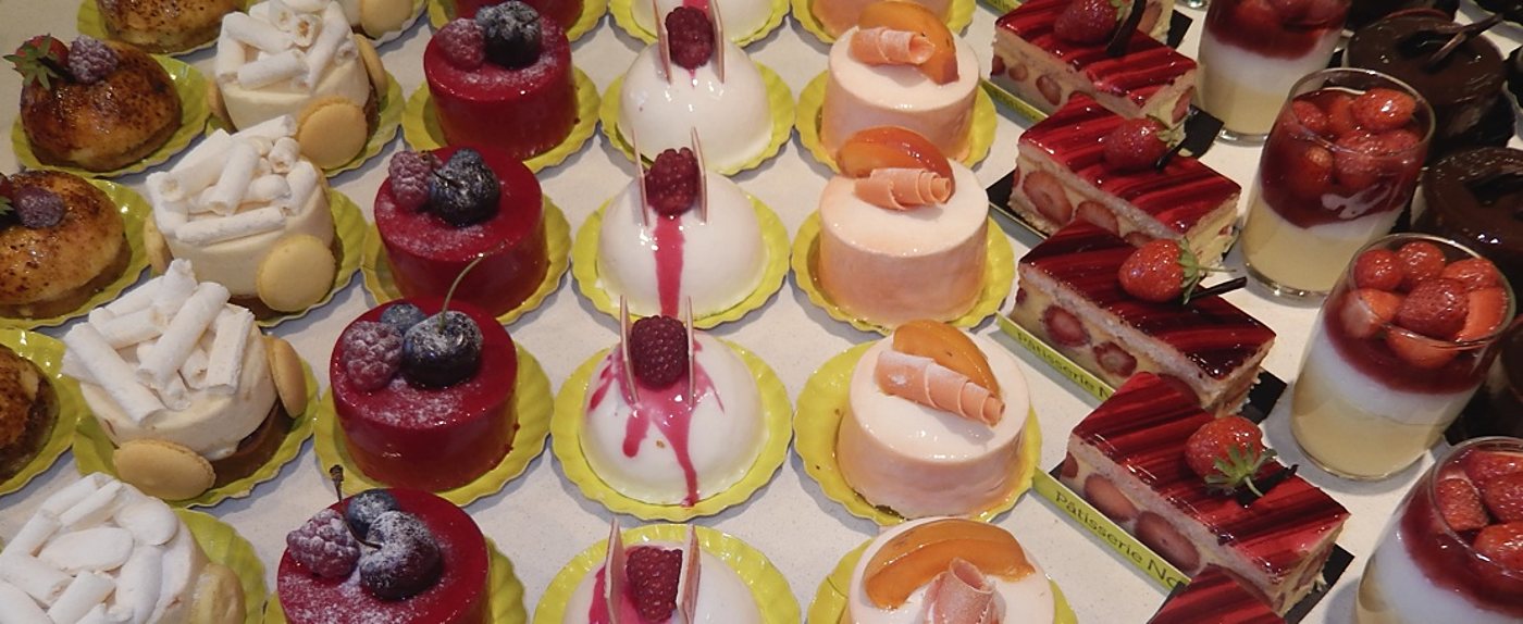 Vue de dessus de rangées de petits gâteaux individuels garnis de framboises, myrtilles, fraises et abricots, posés sur des caissettes jaunes dans une vitrine de pâtisserie.